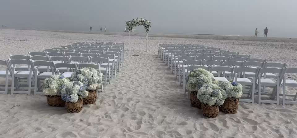 Beach ceremony with a white floral arch and hydrangea baskets along the aisle